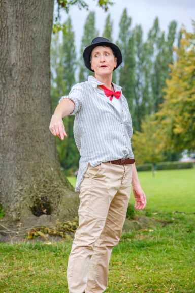 Person in a grey shirt and hat gesturing in a park with trees in the background.